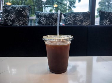 Plastik bardakta buzlu, soğuk kahve, samanla birlikte, siyah yastıklı, rahat bir kafede beyaz bir masada oturuyor.