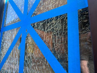 Close up, selective focus on shattered glass taped up with blue tape outside a business on a sunny day