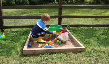 Kızıl saçlı çocuk kum havuzunda oyuncaklarla oynuyor.