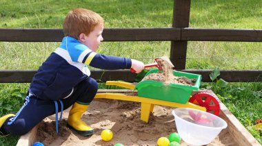 Kızıl saçlı çocuk kum havuzunda oyuncaklarla oynuyor.