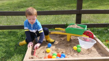 Kızıl saçlı çocuk kum havuzunda oyuncaklarla oynuyor.