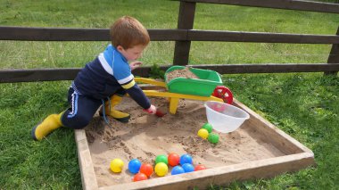 Kızıl saçlı çocuk kum havuzunda oyuncaklarla oynuyor.
