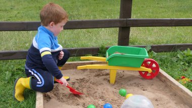 Kızıl saçlı çocuk kum havuzunda oyuncaklarla oynuyor.
