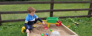 Kızıl saçlı çocuk kum havuzunda oyuncaklarla oynuyor.