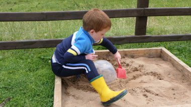 Kızıl saçlı çocuk kum havuzunda oyuncaklarla oynuyor.