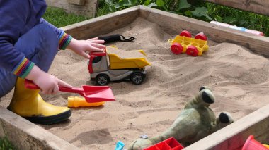 Kızıl saçlı çocuk kum havuzunda oyuncaklarla oynuyor.