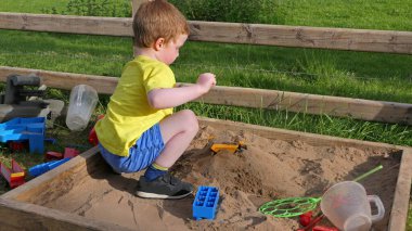 Kızıl saçlı çocuk kum havuzunda oyuncaklarla oynuyor.