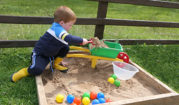Kızıl saçlı çocuk kum havuzunda oyuncaklarla oynuyor.