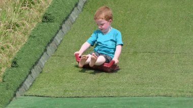 Kırmızı saçlı, mavi tişörtlü çocuk güneşli bir günde bir oyun parkında çimen kaydırağında eğleniyor.