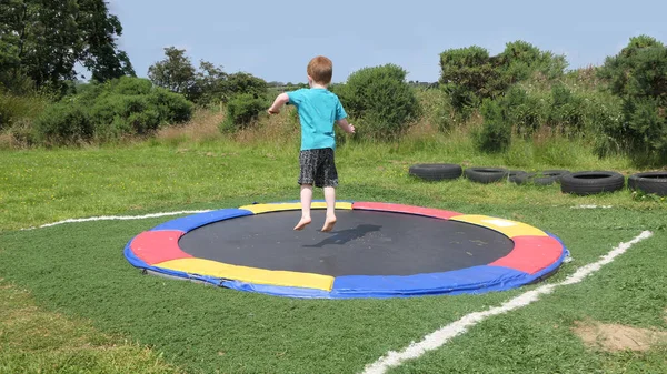 Kırmızı saçlı, mavi tişörtlü, güneşli bir günde bir oyun parkında trambolinde eğlenen çocuk. 