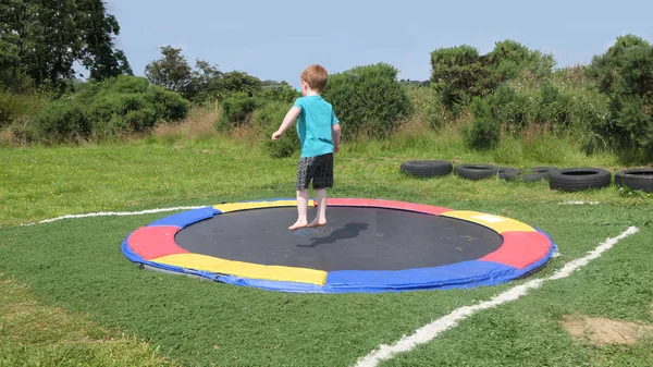 Kırmızı saçlı, mavi tişörtlü, güneşli bir günde bir oyun parkında trambolinde eğlenen çocuk. 