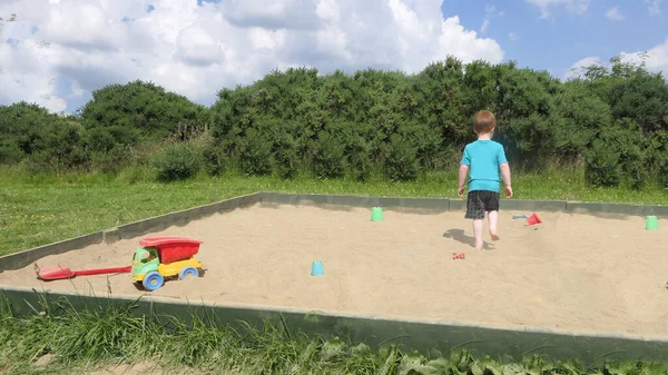 Kızıl saçlı çocuk kum havuzunda oyuncaklarla oynuyor.