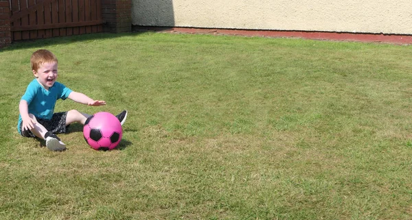 赤い頭の男の子は青いTシャツを着ていて 晴れた日に庭の芝生の上でピンクのサッカーを楽しんでいます — ストック写真