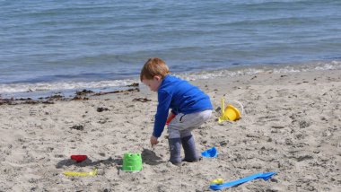 Kızıl saçlı çocuk kumlu bir sahilde kova küreği ve kazıcıyla oynuyor. 