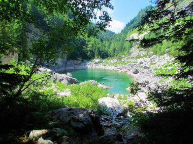Triglav gölleri vadisindeki Kara Göl, Komarca veya Crno Jezero manzarası ve Slovenya 'da güneşli bir günde göl kıyısındaki kayalıklarda duran turistlerin olduğu Julian Alp manzarası