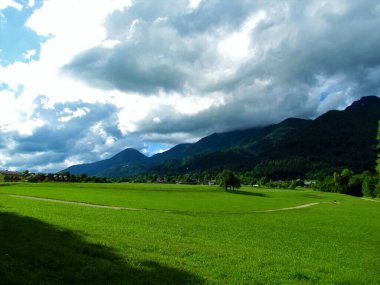 Slovenya 'nın Gorenjska kentinde gökyüzünde bulutlarla kaplı güneş ışığı ve dağlarla aydınlatılmış güzel çayır.