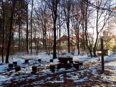 Kışın orman karla kaplıdır. Daire şeklinde ahşap sandalyeler ve ormanın arkasında iskan binası olan bir masa.
