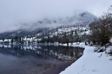 Slovenya 'daki Bohinj Gölü' nün kıyıları karla kaplıydı ve yukarıdaki dağ sisle kaplıydı.