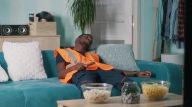 Yorgun bir Afrikalı Amerikalı işçi evde televizyon seyrediyor.