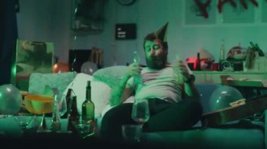 Sarhoş sakallı erkek bira içiyor ve pizza yiyor oturma odasındaki kanepede oturmuş renkli neon ışıkla aydınlanırken dans ediyor.