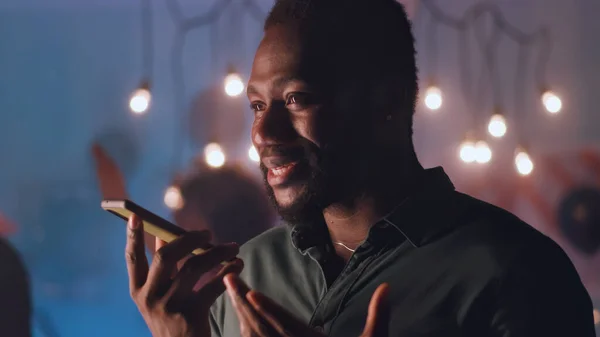 Feliz hombre negro que se comunica a través de mensajes de audio durante la fiesta nocturna —  Fotos de Stock