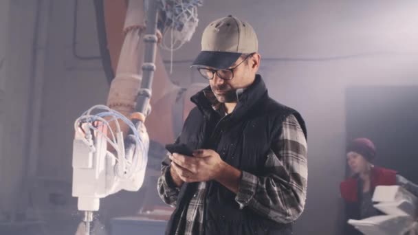 Mężczyzna rzemieślnik za pomocą smartfona w pobliżu robota ramię — Wideo stockowe