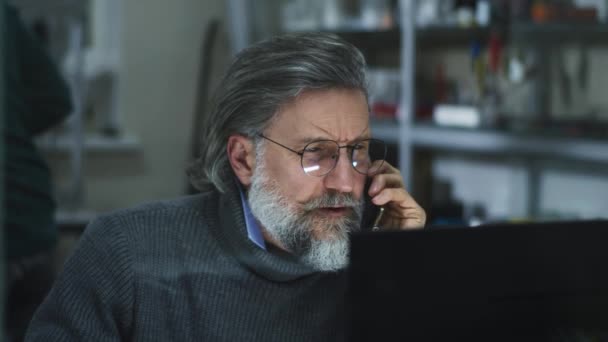 Зрелый дизайнер читает данные с компьютера во время телефонного разговора — стоковое видео