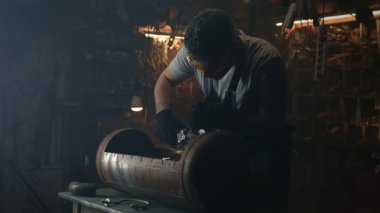 Bir erkek işçi, koruyucu üniforma, gözlük ve eldiven giyerek küçük işletmesi için bir atölyede, bbq ızgarasının detaylarını işliyor.