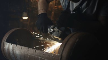 Modern donanımlı loş bir garajda barbekü ızgarası yaparken, iş kıyafetleri ve koruyucu gözlüklü bir adamdan kıvılcımlar yakın çekim.