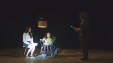 Tekerlekli sandalyede sakat bir aktrisle tiyatro oyuncuları bir oyun provası sırasında spot ışıklarıyla aydınlatılan bir sahnede duygusal roller oynuyorlar.
