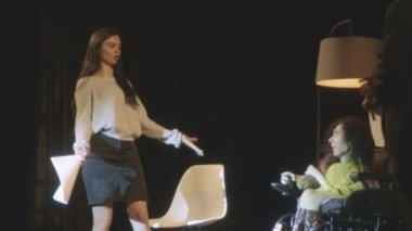 Theater actors with a disabled actress in a wheelchair with scripts rehearsing a scene for a play on a dim theater stage, with decorations and illuminated by a spotlight