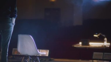 Tiyatro sahnesinin ortasındaki sandalyeye elinde senaryosuyla oturan bir erkek yönetmen sahne ışıklarıyla aydınlanıyor.