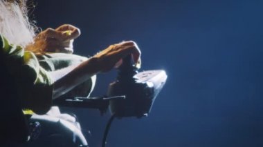 Spinal kas atrofisi olan bir kadın loş bir tiyatro sahnesinde elektrikli sandalye kullanıyor. Sahne ışıkları altında, yönetmenle prova yaparken.