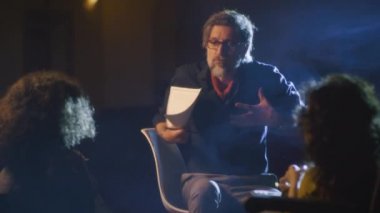 Mature director on a dim theater stage talking to the actor and disabled actress about the performance script and showing how to act more emotionally