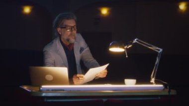 The director in the dark theater reading the script and giving recommendations to the actors, during the rehearsal while sitting at a table with a lamp and a laptop