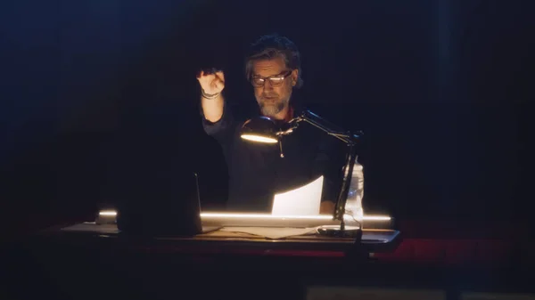 Director in glasses reading the script of the play under the lamp and looking at the camera, while sitting at the table in the dark hall of the theater