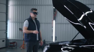 A male mechanic in a virtual reality headset gesturing and standing near the open hood of a car, while making a diagnostics in a car service