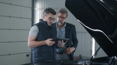 Mechanic watching car diagnostics and maintenance data in a tablet while male client in suit talking to him, in a car service near an opened hood