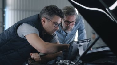 Gözlüklü bir tamirci ve bir araba servisinin kaputunun yanında birlikte duran ve dijital tablet bilgisayar kullanarak araba tanılama sonuçlarını tartışan bir müşteri.