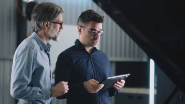 Serious manager in a suit and glasses communicating with a man and diagnosing a car using a gadget in a car service repair shop