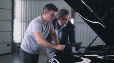Man mechanic talking to client and checking car engine during diagnostics in car service using tablet computer