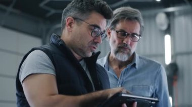 Gözlüklü bir tamirci araba servisinden bir müşteriyle konuşuyor ve araba kontrolü ve tablet üzerinde onarım sonuçlarını gösteriyor.