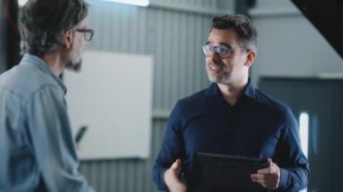 Handheld shot in a repair shop of car service a manager with a tablet greeting a male client talking to him, and consulting about repairing a car after a crash