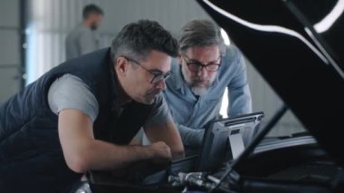 A mechanic or service manager in glasses and a client standing together in repair shop near the opened hood , and discussing the results of car diagnostics using a digital tablet computer