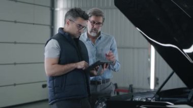 Handheld shot of a car service or mechanic worker in repair shop and a male client standing near the car and watching the data in the tablet, while worker diagnosing the car after a breakdown