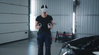 Asian woman car service worker in virtual reality headset with controllers and in uniform standing near the car and doing modern diagnostics
