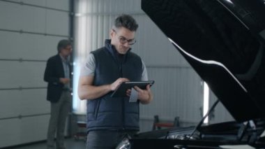 Mechanic in glasses swiping on tablet while standing near the bonnet and checking the results of car diagnostics and repairing in a car service