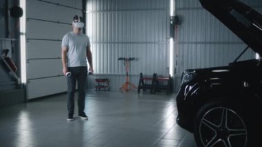 In a car service a man mechanic in virtual reality glasses with controllers moving his hands, while diagnosing and inspecting a car with opened hood