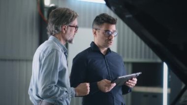 Serious manager in a suit and glasses communicating with a man and diagnosing a car using a gadget in a car service