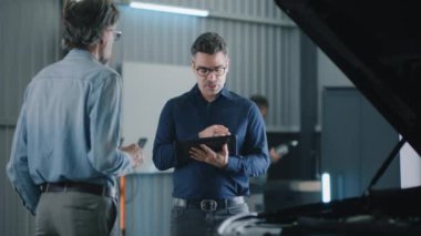 Handheld shot of service manager in repair shop weared casual clothes with a tablet, talking to a male client in a car service and discussing checking and diagnosing a car